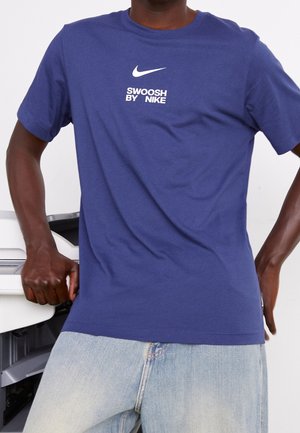 Man in een blauw Nike-T-shirt met de tekst "SWOOSH BY NIKE" leunend tegen een witte printer, gekleed in lichtblauwe jeans.