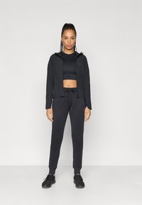 Melnas viegls jaka ar kapuci, apvienota ar melnu īsu augšu un sporta biksēm. Audums ir gluds; dizains ir pieguļošs ar elastīgām aprocēm.