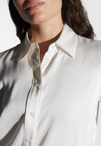 Camicia di seta bianca con chiusura a bottoni, colletto appuntito e superficie liscia con un leggero lucido.