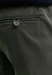 Zielone spodnie z bawełny o teksturowanej powierzchni, z widocznym szarym guzikiem i przeszyciem kieszeni. Wzór logo marki wytłoczony na materiale.