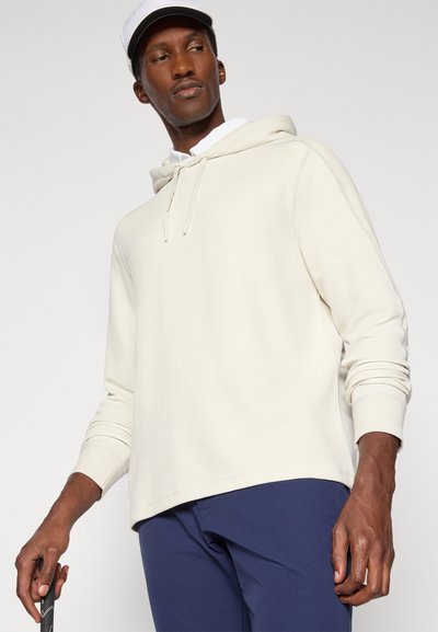 Ljusbeige hoodie med texturerad yta, justerbar dragsko och avslappnad passform, matcher med marinblå byxor och en vit keps.