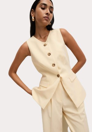 Débardeur sans manches crème avec un décolleté en V profond, cinq boutons décoratifs marron et poches latérales ; porté avec un pantalon sur mesure assorti.