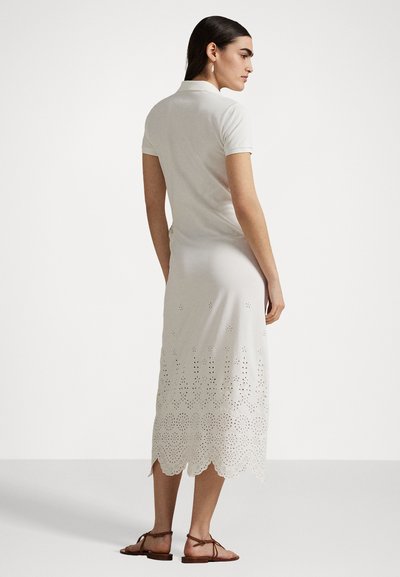 Weißes, knielanges Kleid mit Polokragen und strukturiertem Saum, der mit gewellten Ausschnitten versehen ist. Kombiniert mit braunen flachen Sandalen.