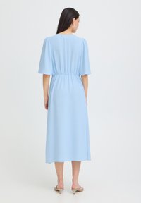 Femme aux longs cheveux foncés portant une robe midi bleu clair à manches courtes et taille froncée, debout devant un fond blanc uni.