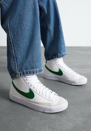 Hvide høje sneakers med grøn Nike-swoosh, båret med blå denimjeans på gråt gulv.