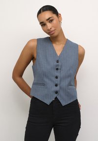 Gilet blu a righe con scollo a V profondo, cinque bottoni neri e due tasche laterali. Indossato con pantaloni neri, dal taglio aderente.