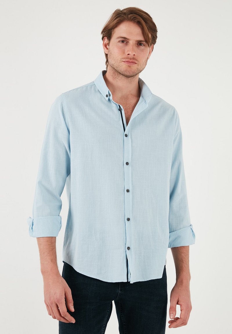 Camisa de manga larga azul claro en una mezcla de lino, con cuello de botón y botones oscuros contrastantes, con puños doblados y un corte relajado.