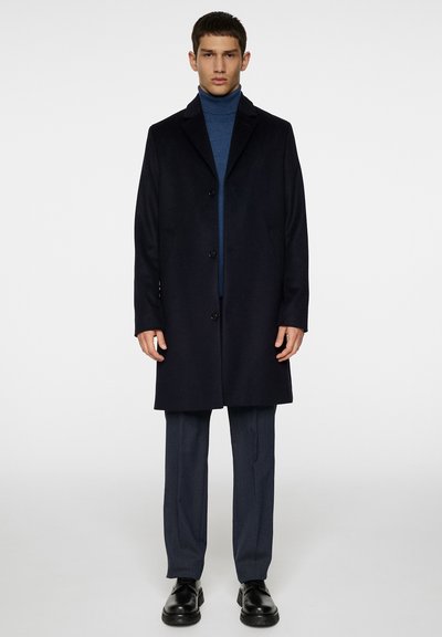 Mann in schwarzem, knöpfbarem Mantel, blauer Rollkragenpullover, grauen Hosen und schwarzen Dressschuhen steht vor einem weißen Hintergrund.