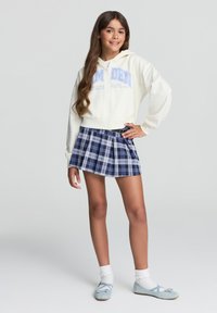 Ragazza in piedi con una mano su i fianchi, che indossa una felpa bianca con zip, una gonna a pieghe a quadratini blu, calze bianche e ballerine azzurro chiaro.