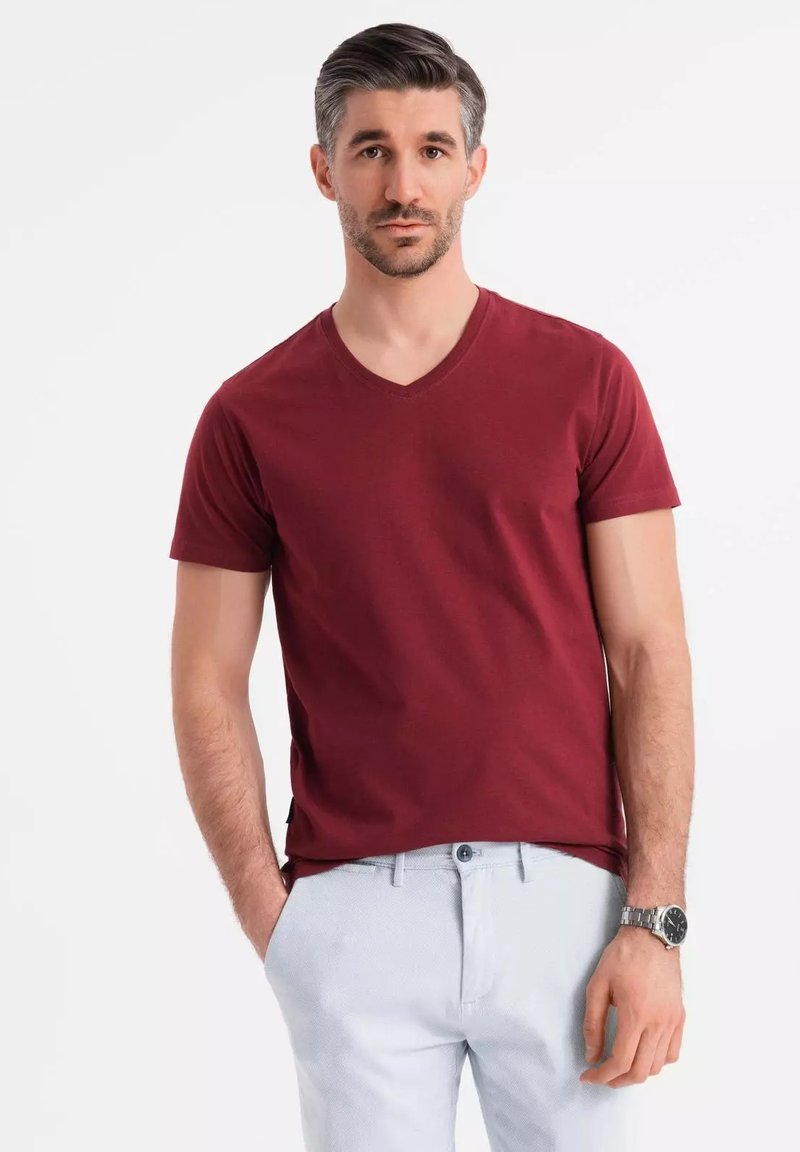 T-shirt a V collo maroon con maniche corte, realizzato in cotone. Il tessuto appare liscio con una vestibilità rilassata. Indossato con pantaloni chiari e un orologio.