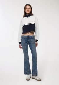 Giovane donna che indossa un pullover corto bianco e blu navy, jeans a zampa d'elefante blu con cuciture frontali e sneakers bianche, in piedi contro uno sfondo semplice.