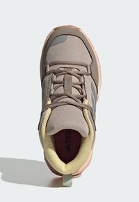 Light brown athletic shoe with a mesh upper, round toe, and textured accents. Features a cushioned collar in pale yellow and a pull tab.