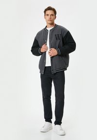 Chaqueta varsity negra y gris con puños acanalados, un logo de "W" en el pecho, sobre una camiseta blanca, combinada con jeans negros y zapatillas blancas.