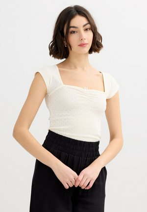 Mujer joven con cabello corto y oscuro, que lleva un top blanco fruncido con cuello cuadrado y pantalones negros de cintura alta, sobre un fondo liso.