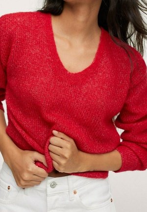 Femme portant un pull rouge à col en V en tissu duveteux et un pantalon blanc, tenant l'ourlet du pull avec les deux mains près de la taille.