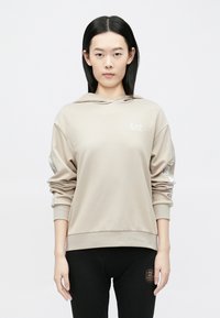 Bēša džemperis ar kapuci un logo priekšpusē, ar aukliņām kapucītī, ribotas aproces un tekstūras detaļas piedurknēs, kombinēts ar melnām biksēm.