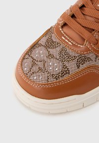 Κοντινό πλάνο στην μύτη ενός καφέ αθλητικού παπουτσιού με μπεζ υφασμάτινο σχέδιο, διακοσμημένο με καφέ και διαυγή στρας, λευκή σόλα και καφέ κορδόνια.