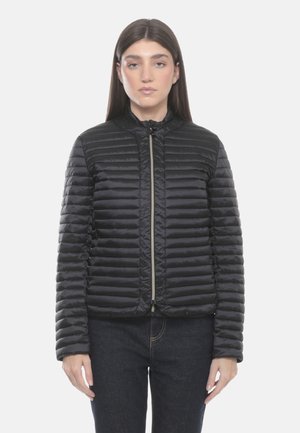 Donna con giacca trapuntata nera con zip e jeans scuri, in piedi davanti a uno sfondo bianco uniforme, rivolta in avanti.