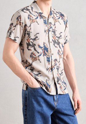Homme portant une chemise beige à manches courtes boutonnée avec un imprimé multicolore de chevaux, légèrement rentrée dans un jean en denim bleu, mains dans les poches.