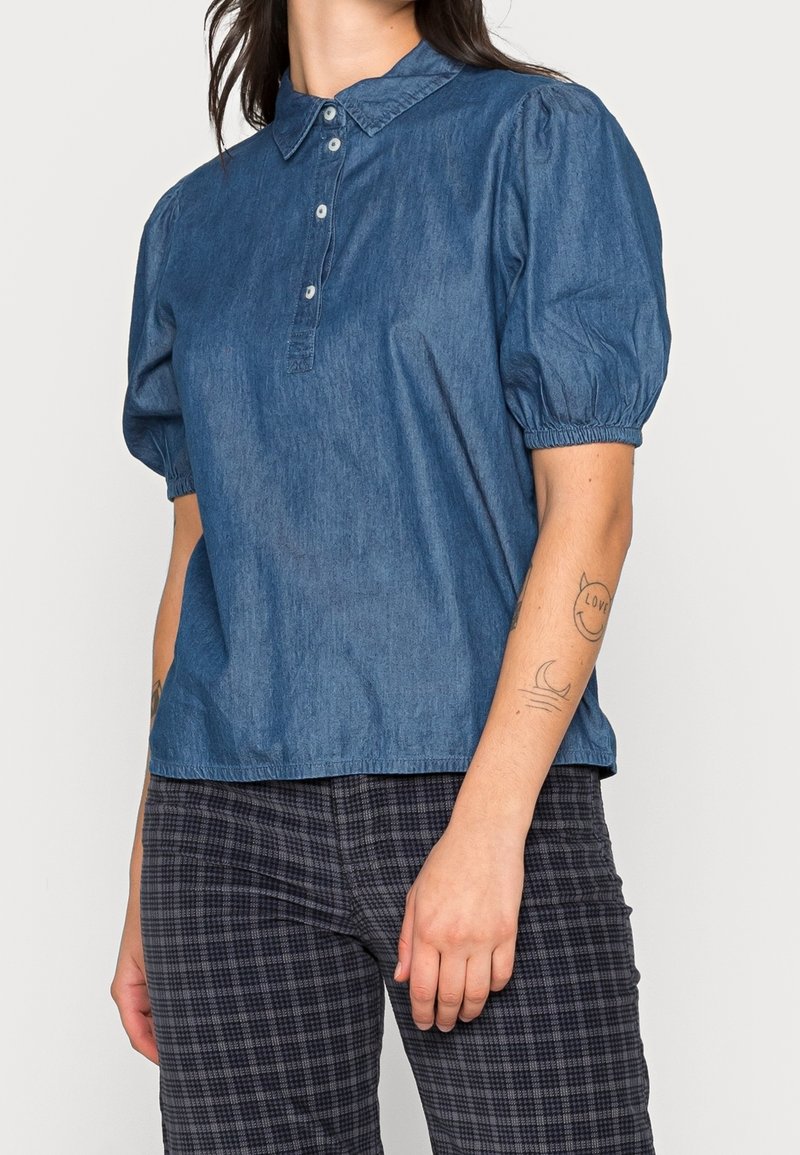 Personne portant une chemise bleue à manches courtes boutonnée avec des manches bouffantes et un pantalon à carreaux noir et gris, montrant des tatouages sur le bras gauche.