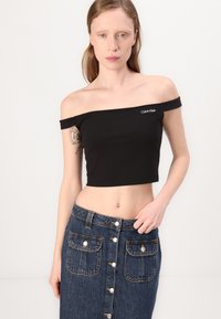 Top corto nero senza spalline con logo Calvin Klein. Abbinato a una gonna di jeans alta in vita con dettagli dei bottoni e tasche frontali.