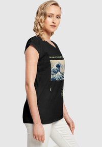 Camiseta negra con gráfico de "La Gran Ola de Kanagawa" en azul y crema, con texto verde abajo. Tejido suave y diseño ajustado.