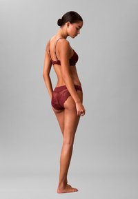 Burgunderfarbener Spitzen-BH mit dünnen Trägern und passendem transparenten Slip, der mit floralen Mustern und weichem Mesh-Gewebe gestaltet ist. Das Modell posiert seitlich.