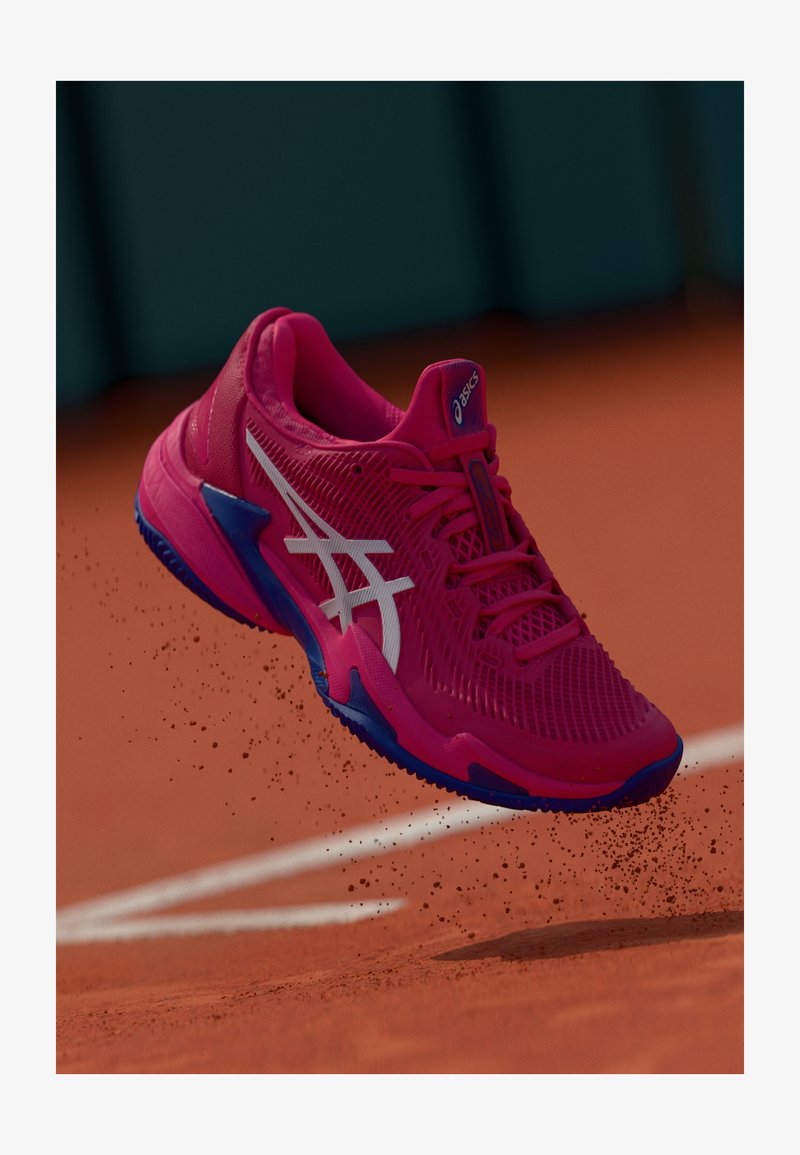 Buty tenisowe Asics mają jasnoróżową siateczkową górę, białe logo, niebieskie akcenty oraz teksturowaną podeszwę o dynamicznym kształcie.