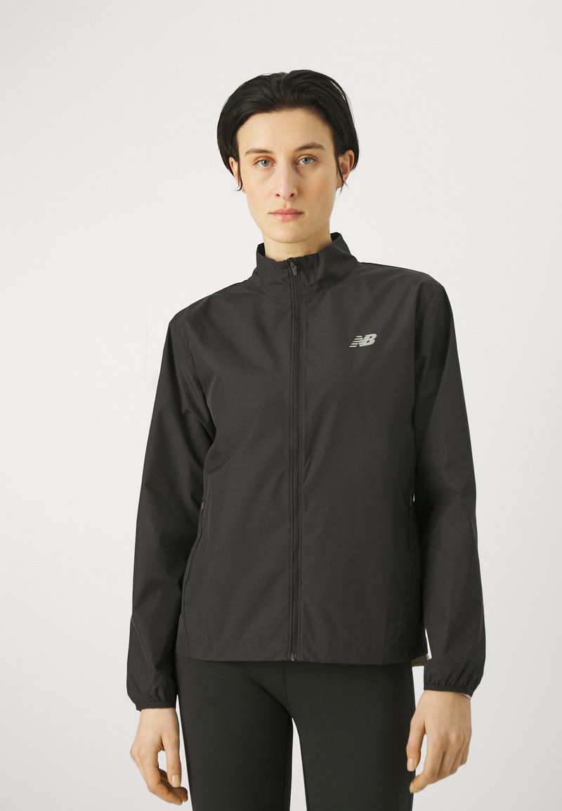Persona che indossa una giacca nera di New Balance con zip e collo alto, insieme a leggings neri, in piedi di fronte a uno sfondo bianco semplice.