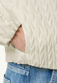 Quilted cream jacket featuring a leaf pattern, with a side pocket and elastic cuff. Worn over light blue denim jeans.