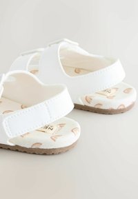 White sandals with a soft fabric lining featuring a rainbow pattern. Adjustable straps and textured brown rubber soles.