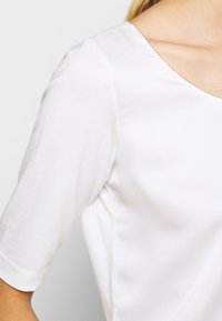 Haut texturé blanc avec des manches courtes et un décolleté large. Le tissu semble léger et doux, avec des coutures subtiles le long du bord.