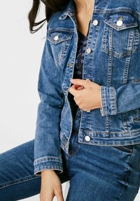 Džínová bunda ve středně modrém odstínu s knoflíkovým zapínáním vpředu, dvěma náprsními kapsami a mírně opranou texturou. Nošená s džínami ve stejné barvě.