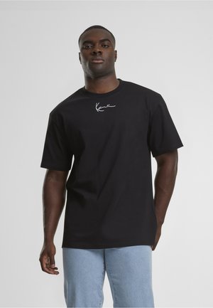 Hombre vestido con una camiseta negra con un logo blanco en el pecho, combinada con vaqueros azul claro, de pie frente a un fondo liso de color gris claro.