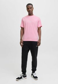 Camiseta de algodón rosa con cuello redondo y mangas cortas, combinada con pantalones jogging negros con un logo. Las zapatillas son negras con detalles en blanco.