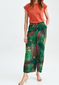 Maglietta rossa in cotone abbinata a pantaloni larghi con stampa tropicale verde, caratterizzati da foglie di palma e accenti di colibrì; include una vita con coulisse.