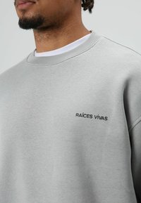 Grijze trui met ronde hals en geribbelde manchetten, met de tekst "RAÍCES VIVAS" in zwarte letters aan de linkerkant. Gladde textuur.