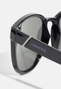 Juodos plastikinės saulės akinių rėmeliai su pilkiais tonais. Yra matinė apdaila ir sidabrinė detalė ant šakės, pažymėta "ARNETTE."