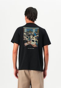 Bărbat îmbrăcat cu un tricou negru ce are un grafic cu un birou aglomerat plin de computere retro și o persoană îmbrăcată în albastru care trece printre birouri.