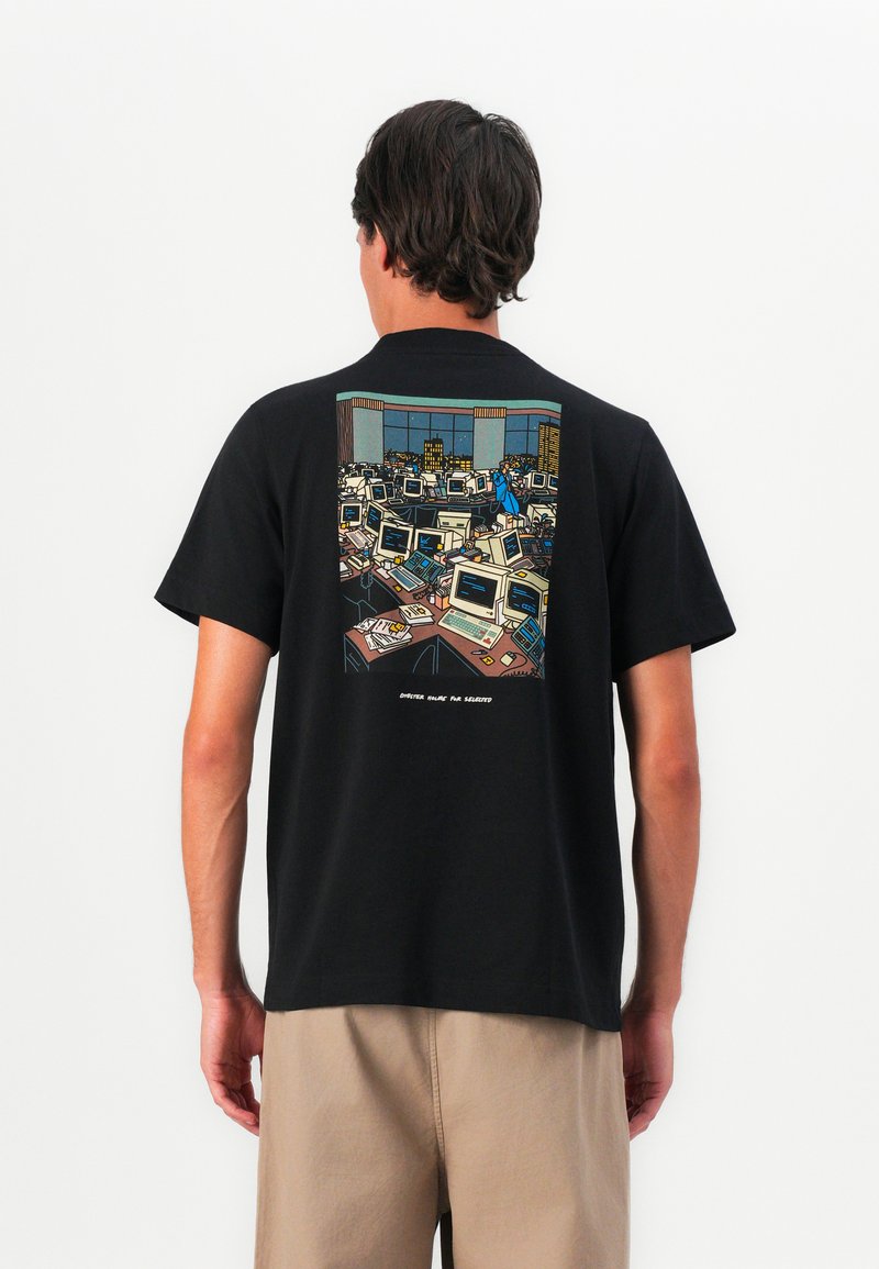 Bărbat îmbrăcat cu un tricou negru ce are un grafic cu un birou aglomerat plin de computere retro și o persoană îmbrăcată în albastru care trece printre birouri.