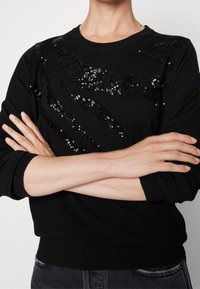 Schwarzer Pullover mit langen Ärmeln und einem Paillettenmuster. Gerippte Bündchen und Saum, Rundhalsausschnitt und glatte Textur.