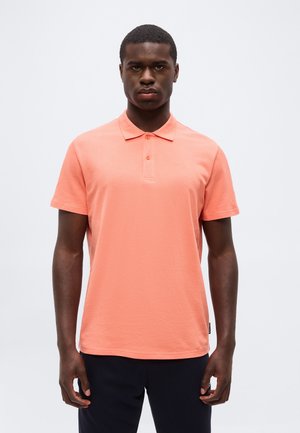 Άνδρας που στέκεται μπροστά φοράει κοντομάνικη μπλούζα πόλο χρώματος σολομού και σκούρο παντελόνι πάνω σε απλό φωτεινό φόντο.