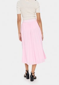 Jupe plissée rose clair avec une large ceinture, en tissu fluide et coupée au niveau du genou, assortie à des sandales à talons noires.