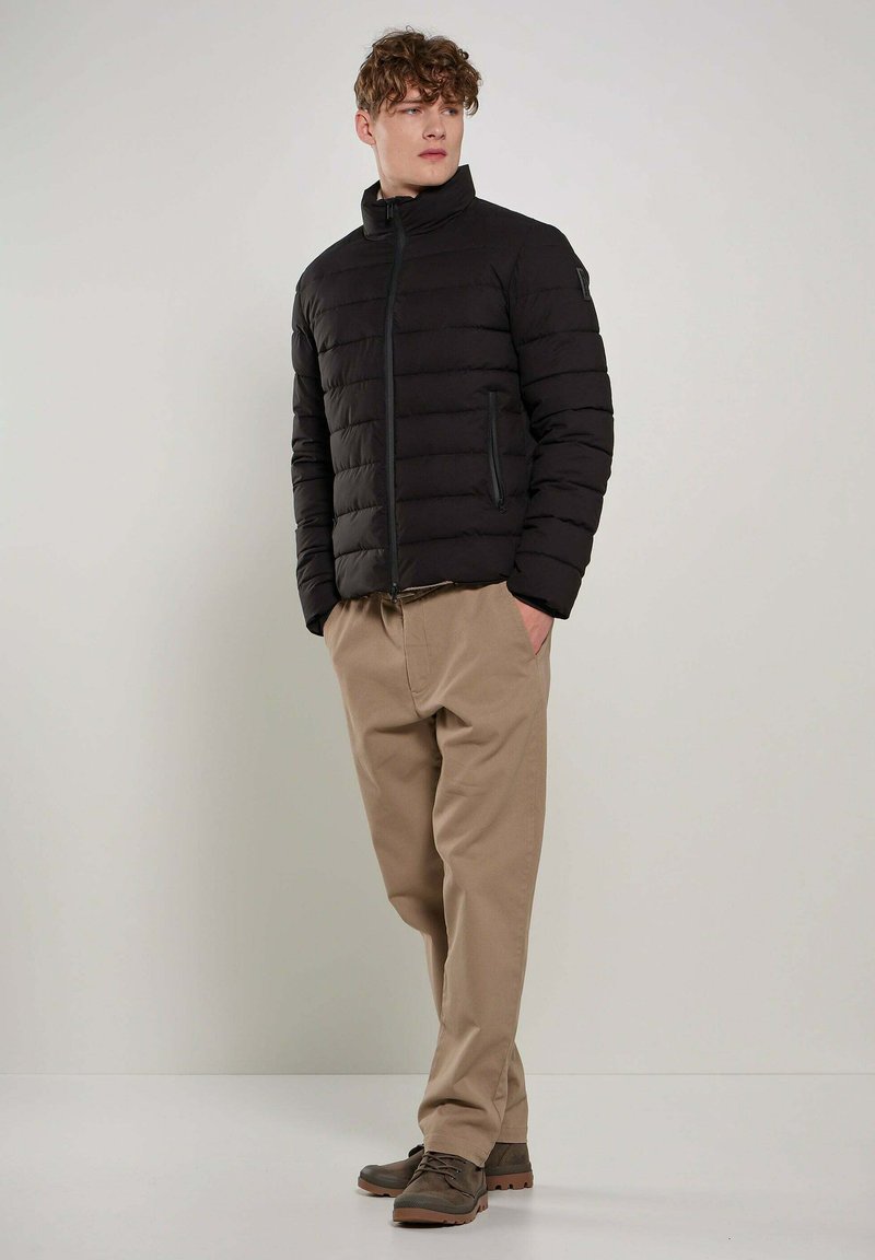 Giacca nera imbottita con colletto alto, trapuntatura orizzontale, due tasche laterali e zip a contrasto. Abbinata a pantaloni beige e scarpe marroni.