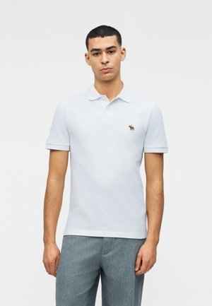 Mężczyzna stojący, ubrany w jasnoniebieską koszulkę polo z niewielkim haftowanym logo łosia na piersi oraz szare spodnie na jednolitym tle.