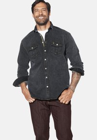 Dunkelblaues Denim-Hemd mit zwei Brusttaschen, hochgekrempelten Ärmeln und kontrastierenden Nähten. Getragen mit dunklen Hosen.