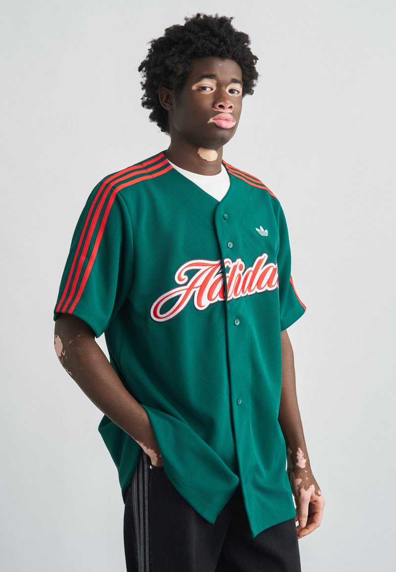 Maillot de baseball vert avec accents rouges, devant boutonné, avec "Athletic" imprimé en blanc. Style avec un pantalon noir à rayures latérales.