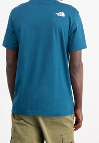 Camiseta de algodón verde azulado con mangas cortas, que presenta un logo blanco en la parte superior de la espalda. Se lleva con pantalones cortos de trabajo beige, con mínimas decoraciones.