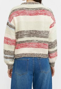 Maglione lavorato a maglia color crema con strisce rosse, marroni e blu. Presenta un design corto con polsini a costine e un collo rotondo. Abbinato a jeans blu.
