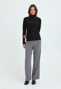 Donna in maglione nero a collo alto, pantaloni grigi a gamba larga e stivaletti neri su sfondo bianco semplice.